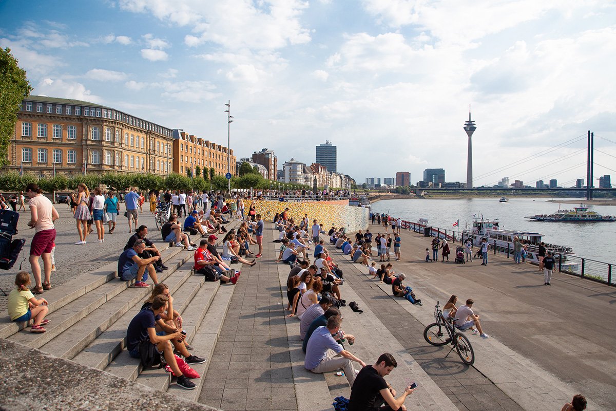 View on the Dusseldorf promenade with Rhein and the Rhein Tower visible in the distance with people sitting on the steps on a sunny day
